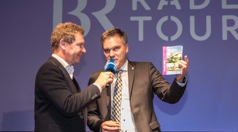 Roman Roell interviewt Wolfgang Bauer auf der BR-Radltour-Bühne. Wolfgang Bauer hält eine zusammengefaltete Karte hoch.