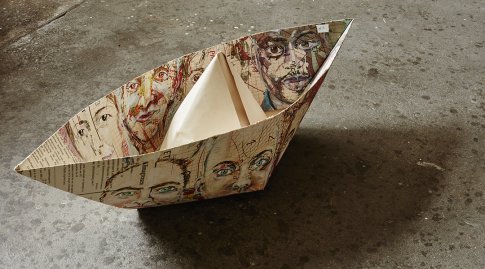 Aus einer alten Schullandkarte gefaltetes Papierschiffchen mit Gesichtsmotiven steht auf dem Boden.