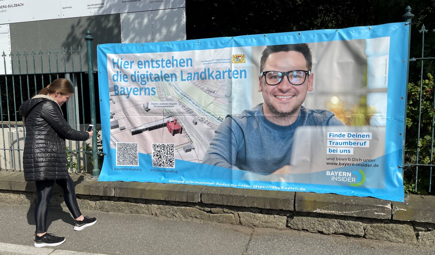 Banner am Bauzaun mit Werbung für den Traumberuf  und einem lachenden jungen Mann, davor eine Dame die den QR-Code scannt