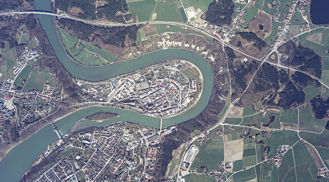 Das Farbluftbild zeigt einen Flussschleife in der sich eine Stadt befindet. Außerhalb der Stadt sind Wälder, Wiesen und Verkehrswege abgebildet.