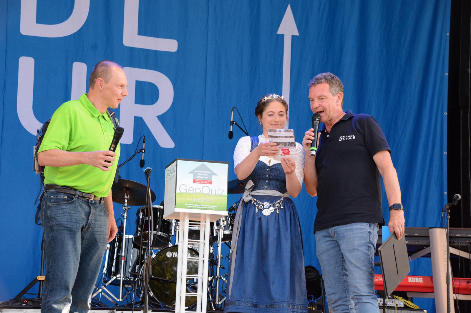 v. l. n. r.: LDBV-Präsident Daniel Kleffel, gemeinsam mit der Bayerischen Bierkönigin Mona Sommer und dem Moderator Roman Roell (BR) auf der BR-Bühne bei der Verkündung der Gewinnerin oder des Gewinners des GeoQuiz