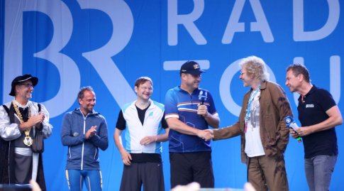 Markus Söder und Thomas Gottschalk schütteln sich umgeben von vier weiteren Männern auf der BR-Bühne die Hände.