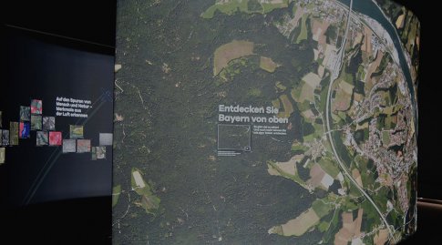 Auf einer gewölbten, wandhohen, beleuchteten Luftbildaufnahme ist ein Ausschnitt Bayerns zu sehen.