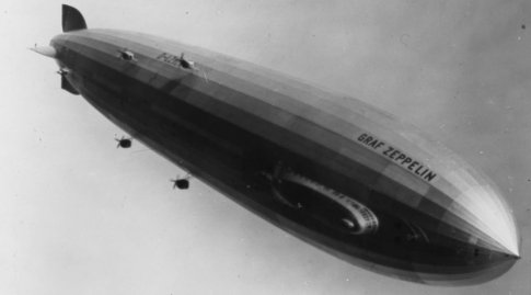Schwarzweiß-Bild eines fliegenden Zeppelins. Auf dem Zeppelin steht 