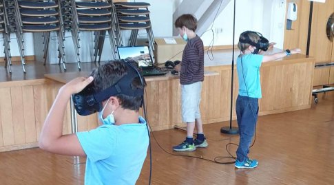 Zwei Jungen sehen stehend in einem Vortragsraum durch VR-Brillen. Dahinter steht ein weiterer Junge und klickt an einem Laptop.