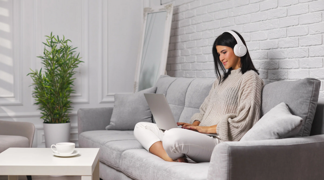 Behördengänge von zu Hause aus erledigen Eine Frau mit dunklen glatten Haaren sitzt mit untergeschlagenen Beinen auf einem Sofa. Sie hat Kopfhörer auf, arbeitet am Laptop und lächelt